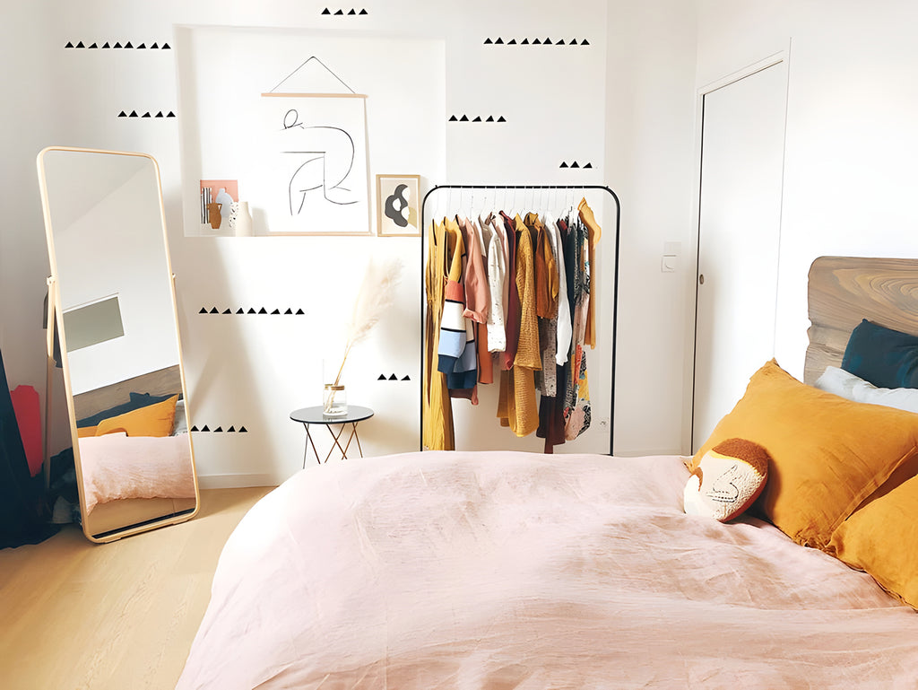 Modern bedroom bathed in natural light, featuring a cozy bed with pink bedding, a full-length mirror, and a clothing rack. ‘Tiny Triangles’ wall decals add a chic accent to the room.