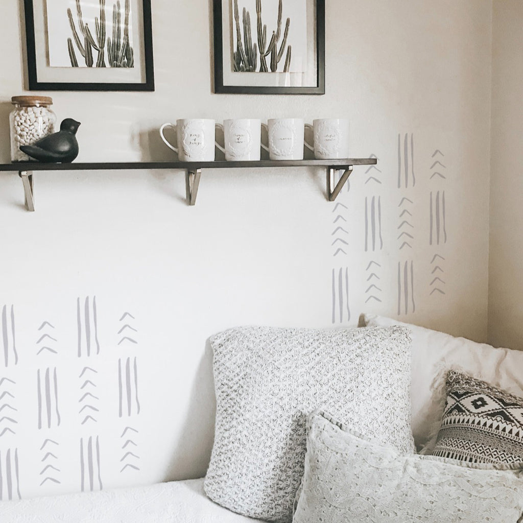 A cozy room corner with a white sofa adorned by a patterned cushion. Above, a floating shelf displays two framed artworks, a decorative bird figurine, and four hanging mugs. The wall features Tribal Geometric Lines, Wall Decals, adding an artistic touch to the space.