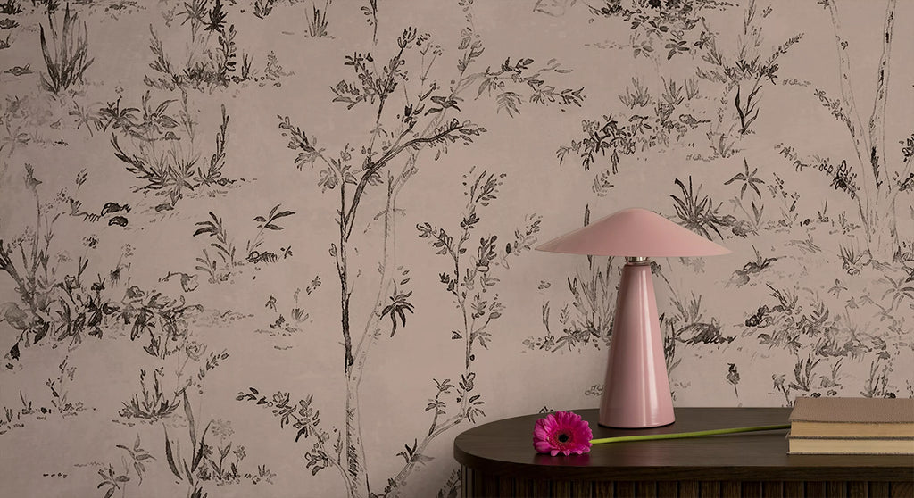 Trisha Floral Pattern Wallpaper, in a subtle nude shade, is featured on a wall of a room. The room is furnished with a brown table, which is adorned with a pink lamp, a pair of stacked books, and a flower.