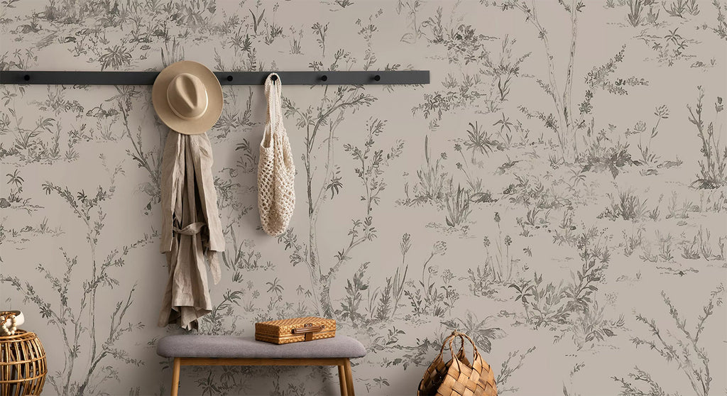 Trisha Floral Pattern Wallpaper, in a sand color, is showcased on a wall of a room. The wall serves as a backdrop for a jacket and a hat hanging alongside a woven bag. Nearby, there is a chair accompanied by a rattan case and basket.