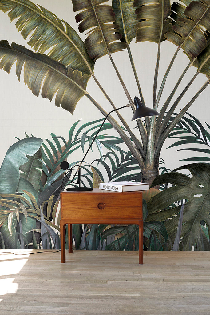 The Tropical Forest Mural Wallpaper, featuring a multicolor, adorns the wall of a room furnished with a vintage wooden table. On top of the table, there is a white book and a black lamp