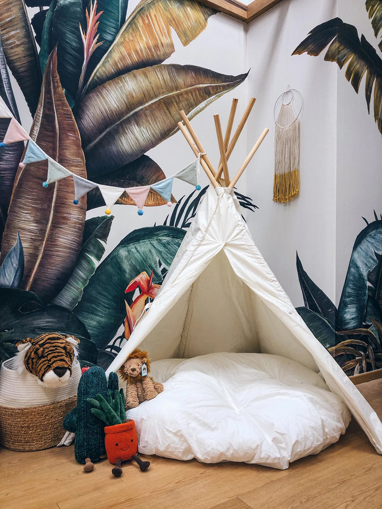 The Tropical Forest Mural Wallpaper, in Multicolor, is displayed on a wall in a kid’s playroom. The room is playfully decorated with a tent and is scattered with several plush toys