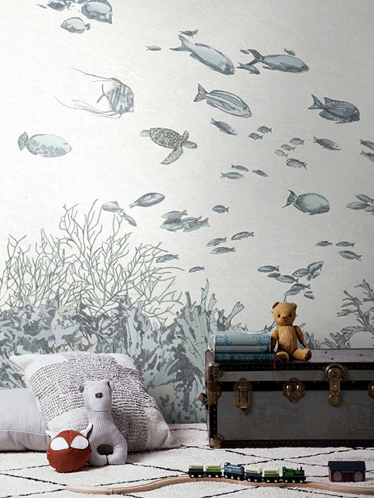 Under the Sea, Wallpaper in dusk blue sets the theme in a room that features a grey treasure chest with  a brown stuffed toy, and other stuffed toys are comfortably nestled on the pillow. Train toys can be seen scattered on the floor, adding to the room’s playful charm.