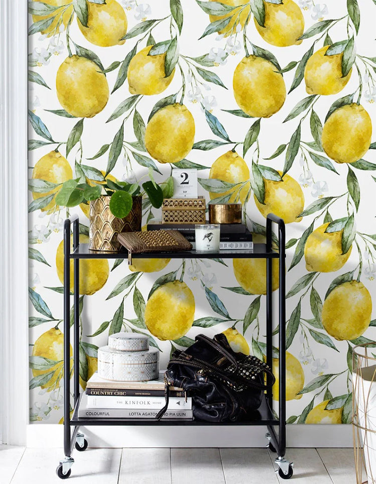 A cozy corner of a room featuring a two-tiered black metal cart filled with books, decorative items, and a black bag, set against a vibrant ‘Vintage Lemons, Pattern Wallpaper.