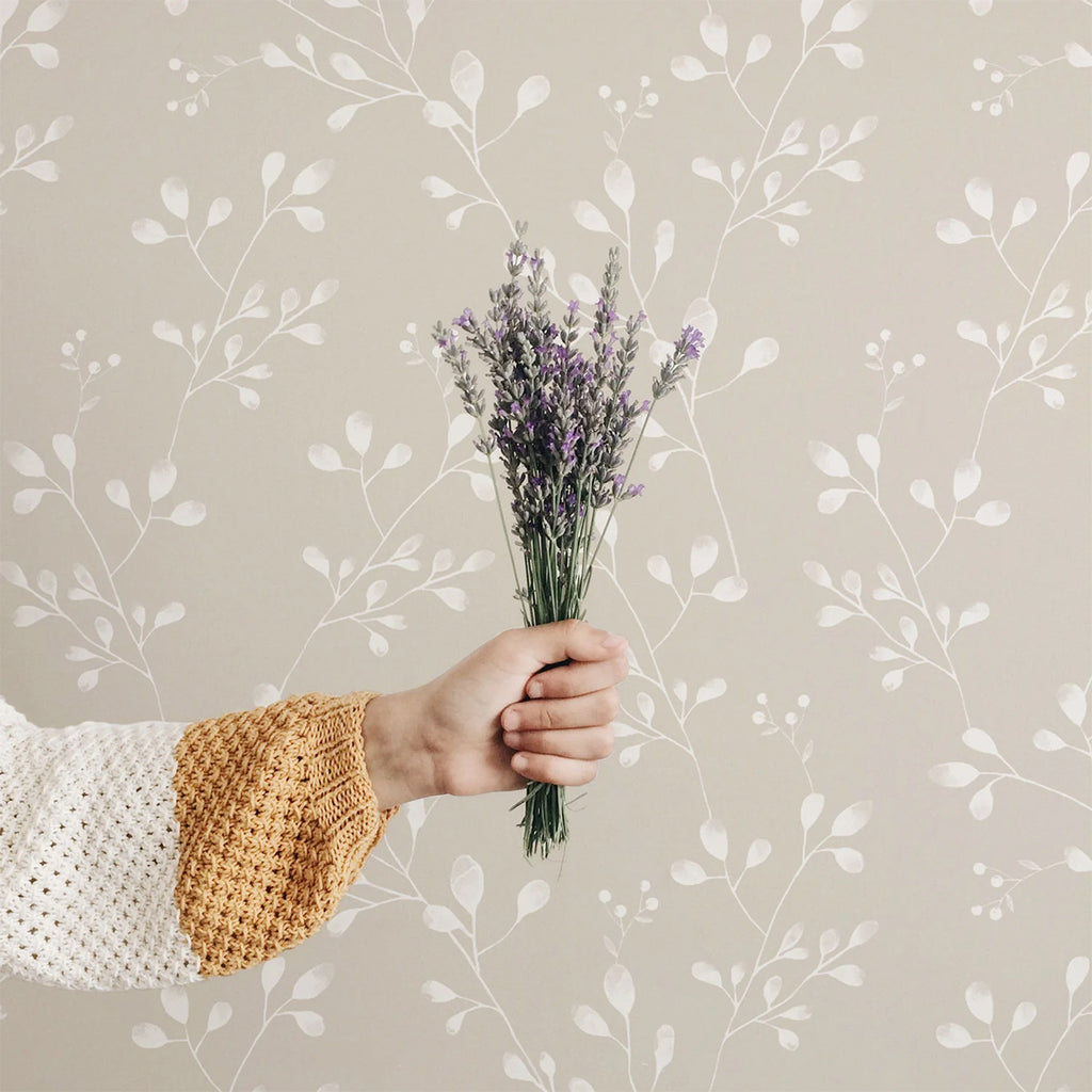 A hand in a mustard yellow knitted sweater holds a bouquet of lavender against a serene backdrop of Vintage Wildflower pattern wallpaper, creating an aesthetic visual.