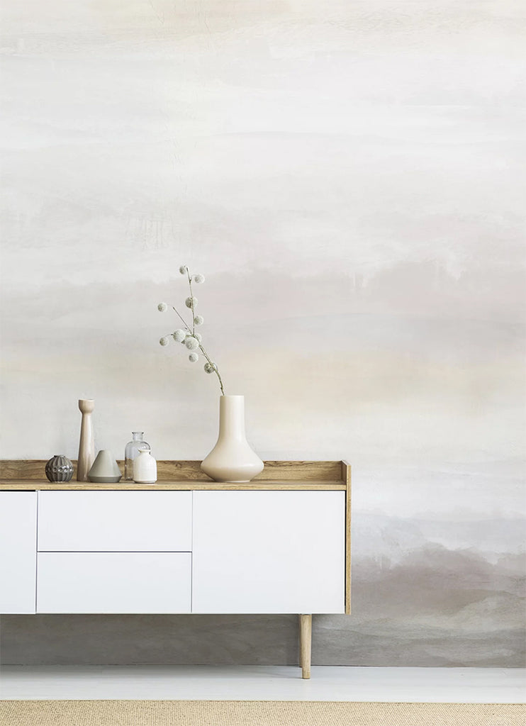 In a room, a wall features the Vista Watercolor Ombre Wallpaper in Sand, creating a striking contrast with a white table. The table is adorned with multiple ceramic vases, adding to the room’s aesthetic appeal.