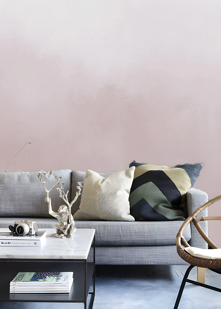 A Blush Pink Watercolour Gradient Ombre Mural Wallpaper is featured on a wall, complemented by a grey sofa adorned with variously designed pillows. Adjacent to the couch, a marble-textured coffee table and a rattan chair complete the setting.