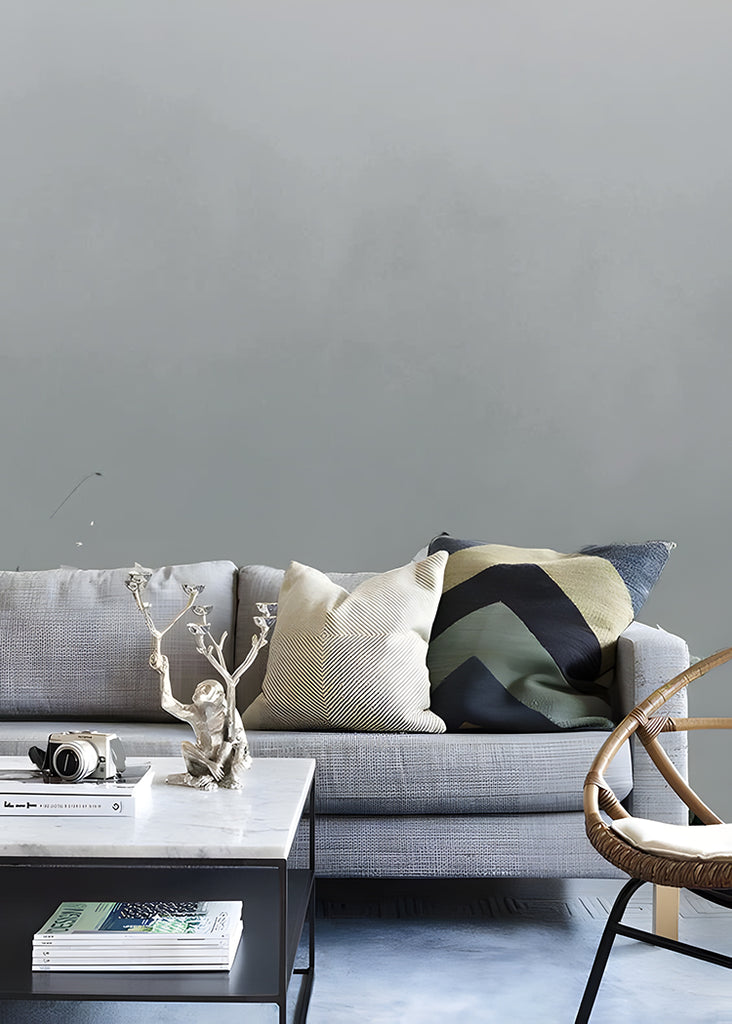 A grey Watercolour Gradient Ombre Mural Wallpaper is featured on a wall, complemented by a grey sofa adorned with variously designed pillows. Adjacent to the couch, a marble-textured coffee table and a rattan chair complete the setting.