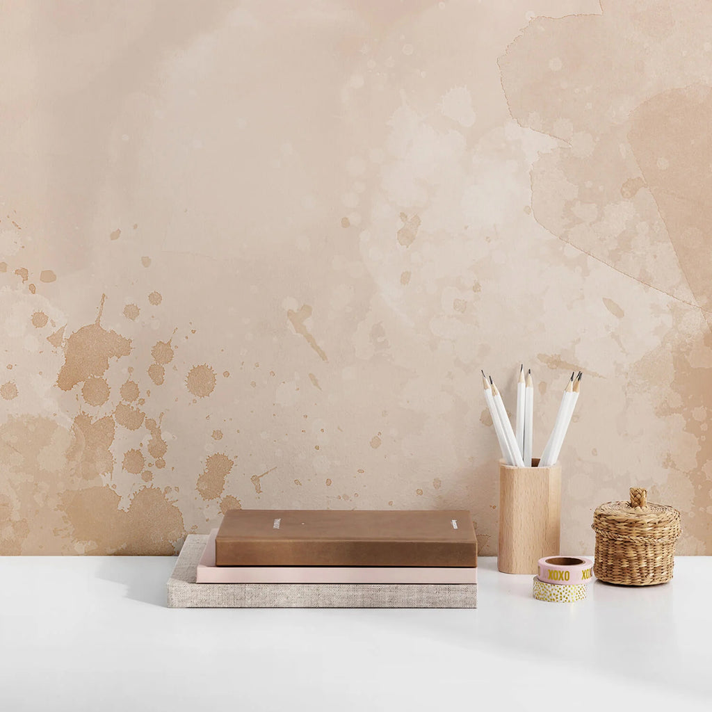 A serene room featuring Watercolour Pastel, Ombre Mural Wallpaper with soft beige and brown tones. A wooden tray, a container of white brushes, a woven basket, and a cup with text are on a surface