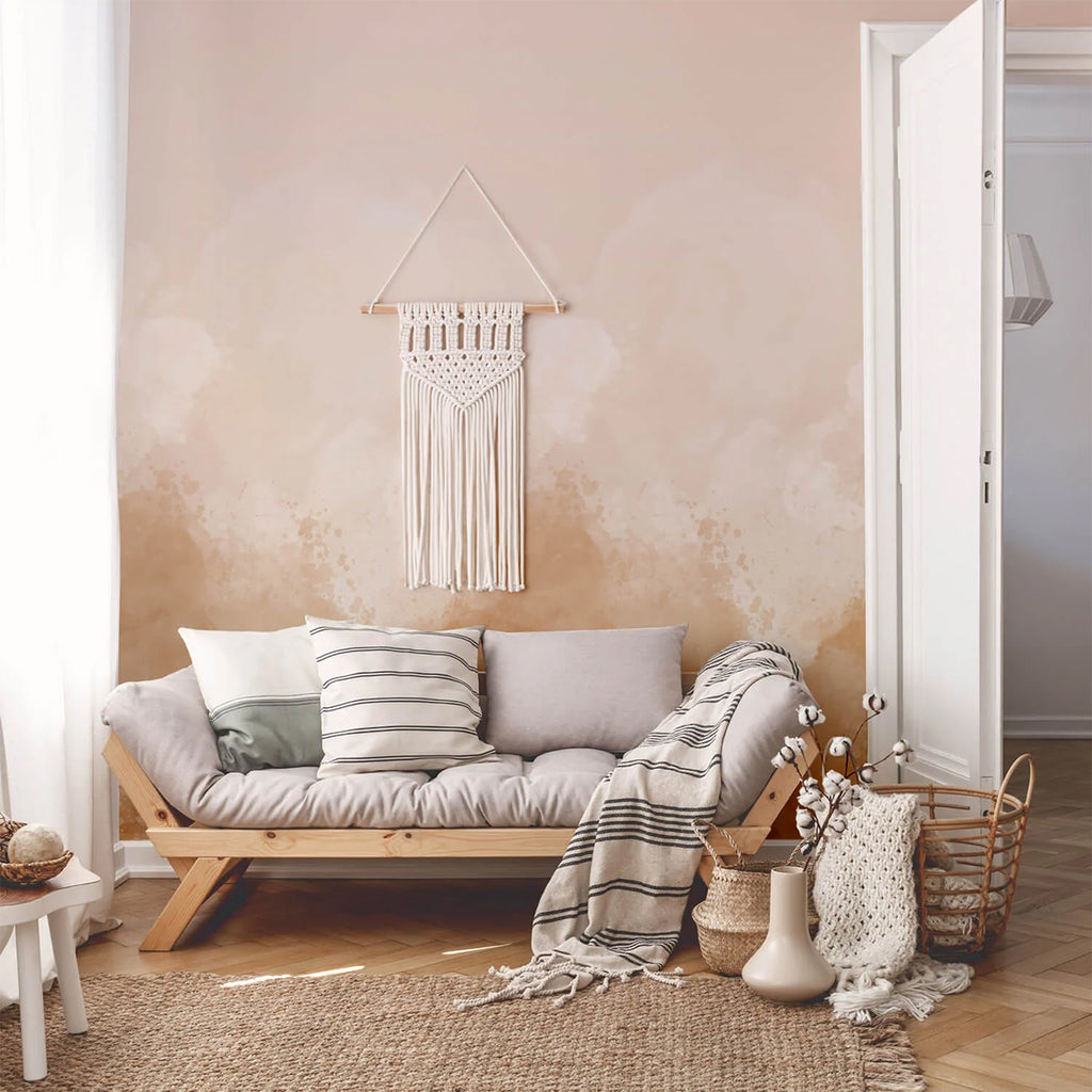 A cozy room with Watercolour Pastel, Ombre Mural Wallpaper. Features include a wooden sofa with cushions, a macrame wall hanging, and a small wooden table with a decorative bowl, creating a warm space.