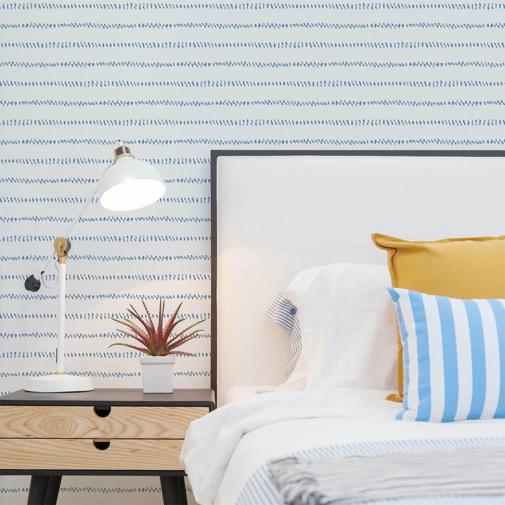 Cozy bedroom scene with a modern bed against a ‘Watercolour Spot Lines’ wallpaper. The bed has white bedding, a striped blue and white pillow, and a yellow pillow. A mounted white lamp illuminates the space.
