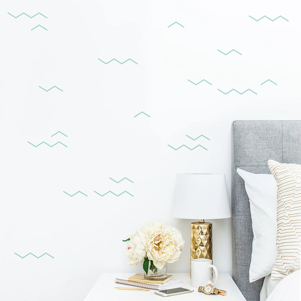 A cozy corner of a room featuring a bed with a gray upholstered headboard and white bedding. A white nightstand holds a stylish lamp, fresh flowers, and an open book. The room is enhanced by a wall adorned with Waves, Blue Pattern Wallpaper.