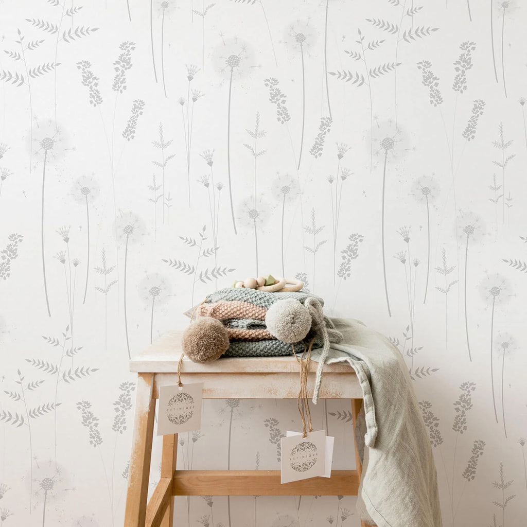 Willow Flower, Grey Pattern Wallpaper adorns a room’s wall, featuring delicate grey floral designs. A wooden stool with folded textiles and tags rests against the wall, under soft, natural light.