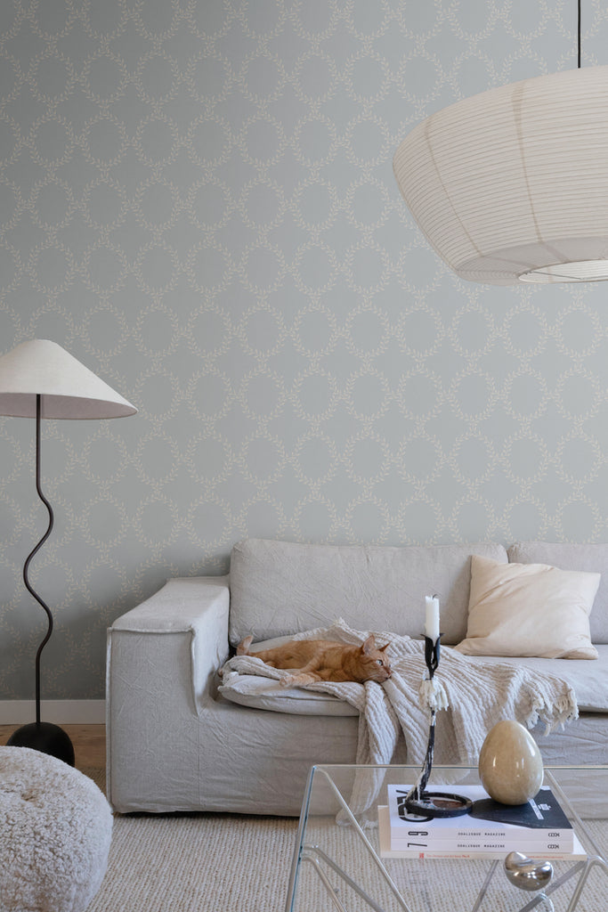 Wilma Wreath Patterned Wallpaper in Dusk Blue Featured on a wall of a living area with a grey sofa with a dirty white fabric and a pillow, along with a glass coffee table, that has a candle and a marble ornament on top, and a lampstand beside it. 