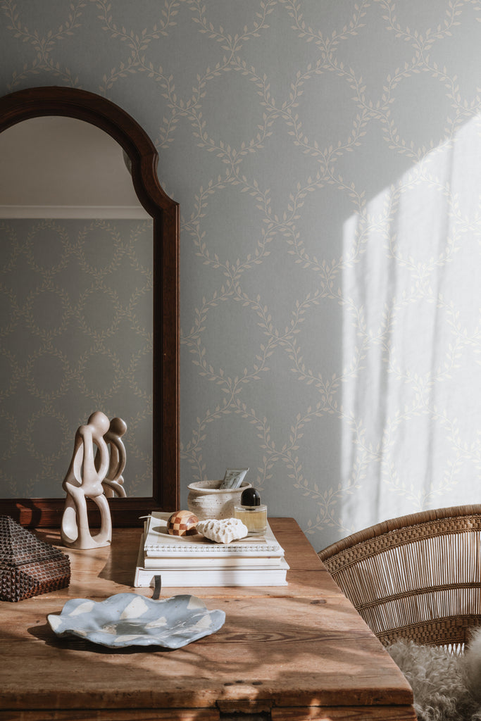 Wilma Wreath Patterned Wallpaper in Dusk Blue Featured on a wall of room with a wooden table, and has books, a mirror, and an abstract sculpture on top, and a rattan chair