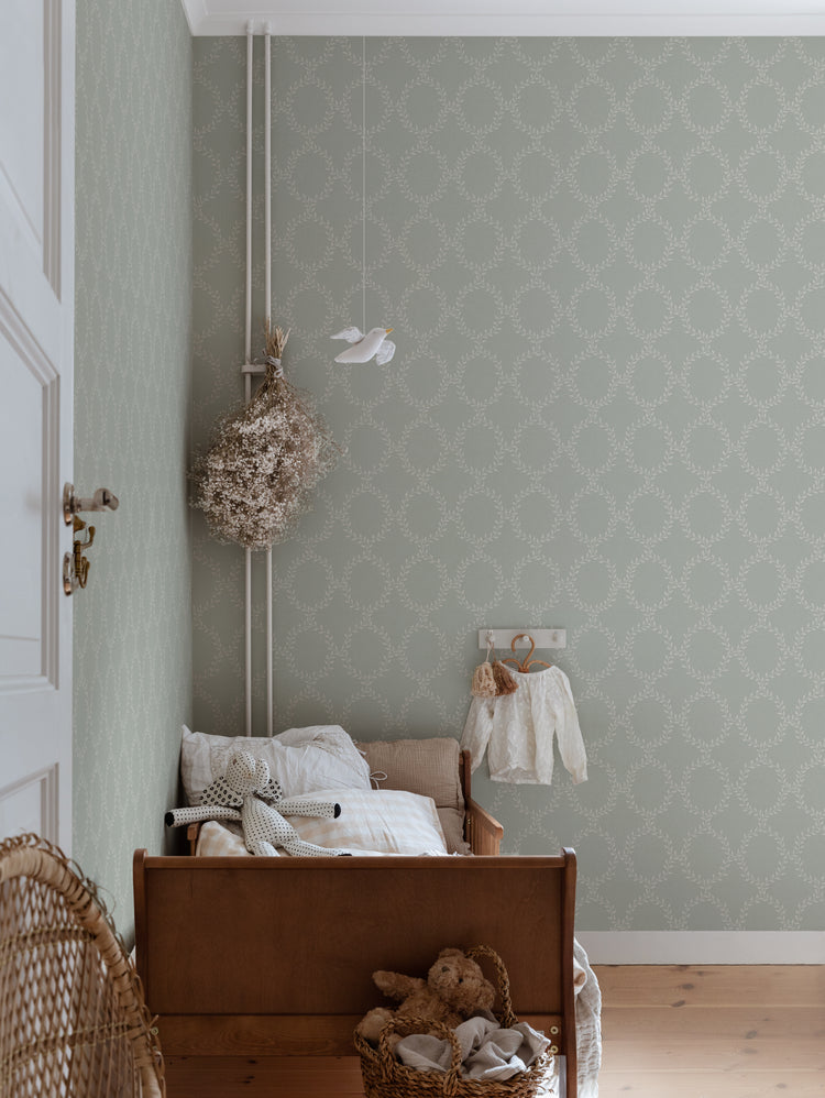 Wilma Wreath Patterned Wallpaper in Sage Featured on a wall of a kid’s bedroom with a small bed in a wooden frame, a basket with a stuffed toy and a rattan chair
