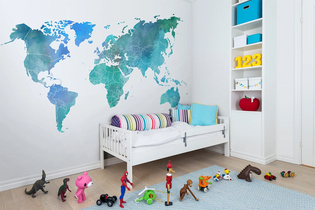 Your Own World Map, a mural wallpaper in emerald green/blue, is featured in a kid’s room. The room is furnished with a crib that has a multicolored bolster pillow, and it is surrounded by a variety of toys.
