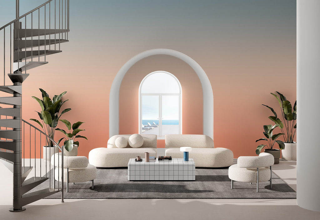 A serene living room with a large arched window revealing a calming seascape. The space features a white, plush modular sofa adorned with round cushions, flanked by potted plants. A spiral staircase adds an architectural touch to the left. The walls showcase the Luminate Gradient, Ombre Mural Wallpaper in Dawn, transitioning colors.