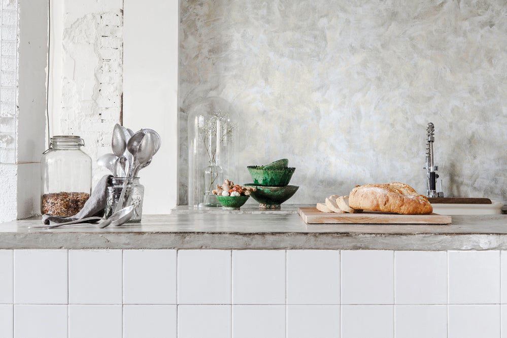 Polished Concrete wallpaper enhances the ambiance of a room featuring a table adorned with a jar of water and plants, along with ceramics used for kitchenware.