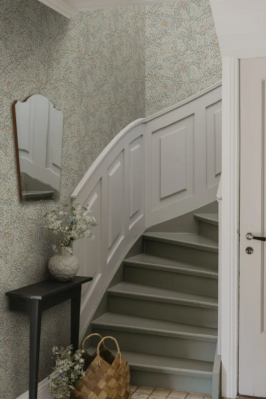 Botanical staircase wallpaper in soft sage and grey above a white panelled curved staircase with a wall mirror and black console table