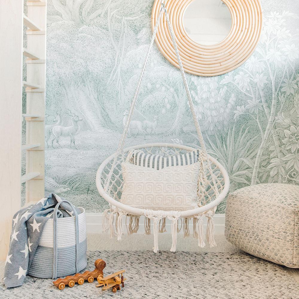 Jungle Land wallpaper with soft hazy jungle trees behind a hanging rattan chair and knitted pouf in a reading corner